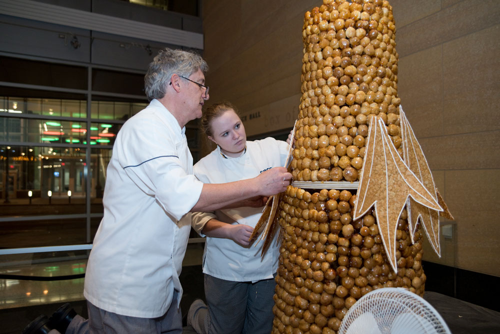 Professor Gilles Renusson, a certified master pastry chef and renowned sugar artist, attaches a branch to the croquembouche, made of 1400 shous (unfilled cream puffs). Student Isabella Patterson assists. It took 24 lbs of caramel to glue together