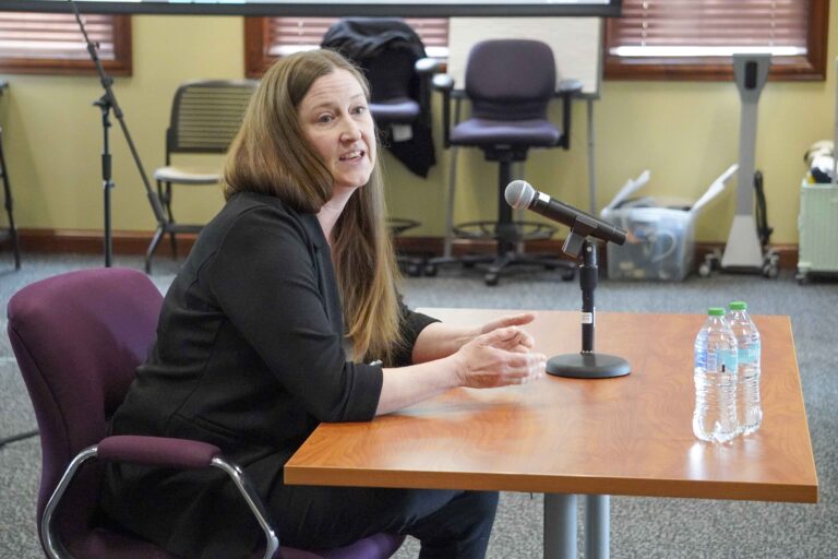 A woman sits at a table, in front of a microphone and bottles of water