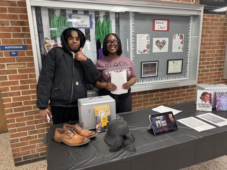 A high school student and youth advocate stand behind a Black History Month exhibit table
