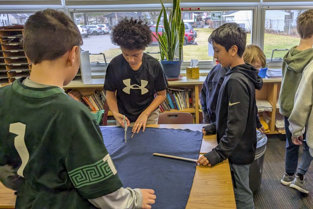 Three court-grade children measure a square of fabric on a table