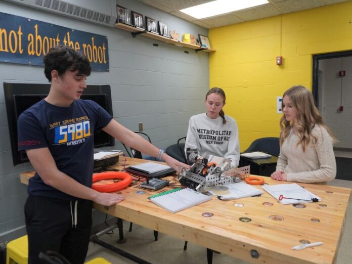 Three students gesture to a robotics team robot on a table